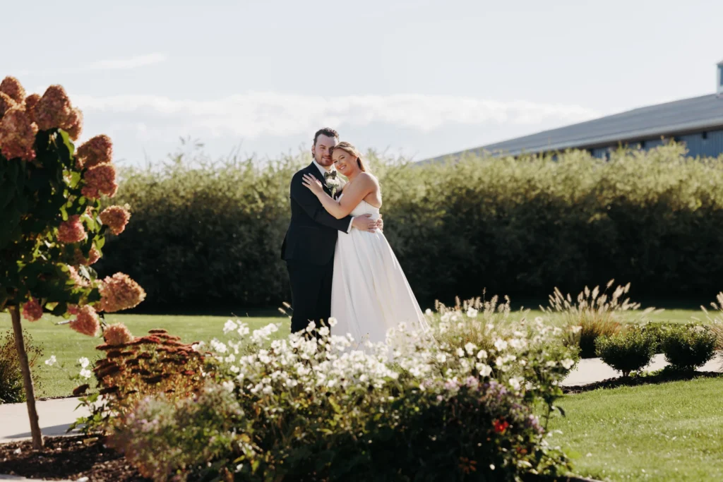 Bride and groom leaning into each other looking at the camera with a garden in the foreground on a sunny day for a fall wedding in Rochester, New York at Pomona at Blue Barn captured by Wolf Cry Productions