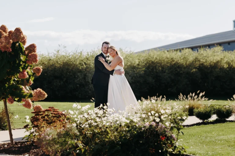 Bride and groom leaning into each other looking at the camera with a garden in the foreground on a sunny day for a fall wedding in Rochester, New York at Pomona at Blue Barn captured by Wolf Cry Productions